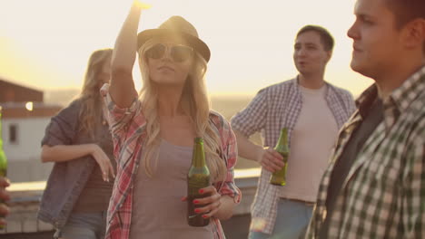 A-woman-in-glasses-and-a-hat-moves-in-a-dance-with-her-two-girlfriends-and-three-boyfriends-on-the-roof.-She-smiles-and-enjoys-the-time-with-beer-in-a-summer-evening.